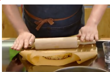 Eine Person mit einer dunkelblauen Schürze, deren Gesicht man nicht sieht, rollt etwas mit einem Nudelholz zwischen zwei Stücken Backpapier aus.