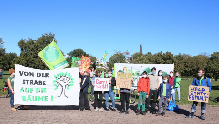 Eine Gruppe von Jugendlichen und jungen Erwachsenen steht vor einem Park. Sie halten Banner und Pappschilder, auf denen steht: Von den Straßen auf die Bäume! Trees for Future. Danni bleibt! Aufbäumen gegen Abholzung. Wald statt Asphalt. Klima retten!