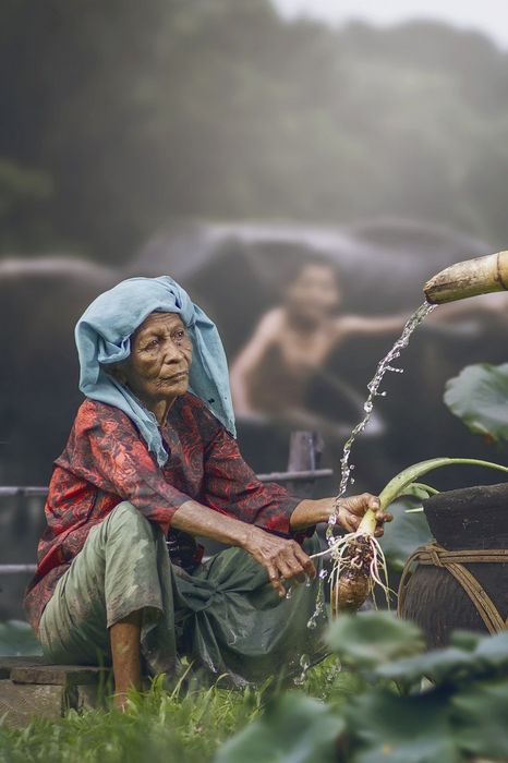 Eine asiatische Frau sitzt vor einem Rohr, aus dem Wasser kommt und wäscht Gemüse.