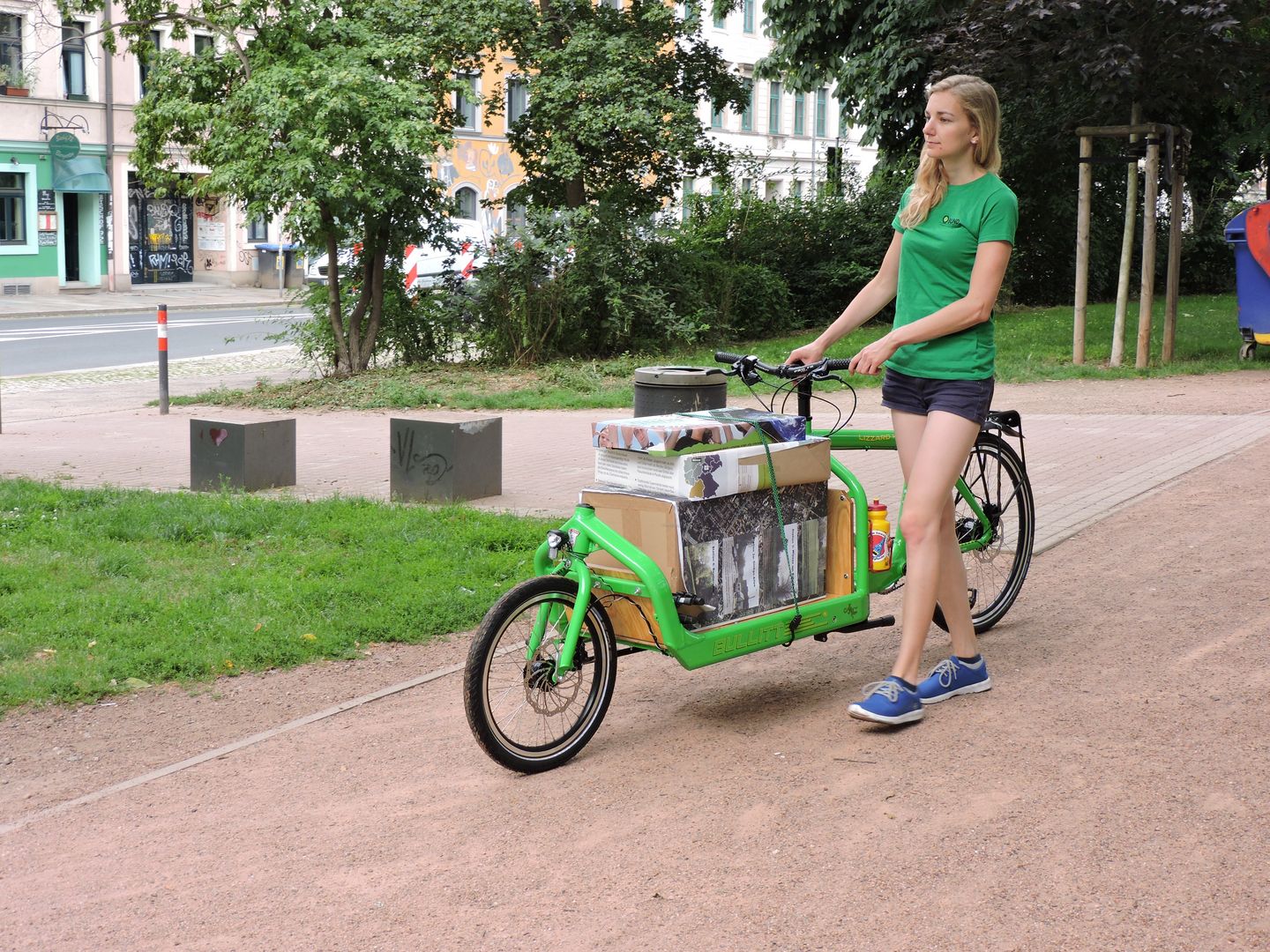 Frau schiebt grünes Lastenrad mit Kartons auf der Ladefläche durch den Alaunpark.