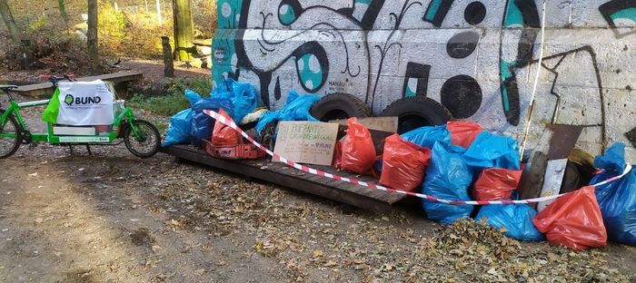 Viele gefüllte Müllsäcke stehen an einer mit Graffitti besprühten Mauer. Links daneben das grüne Lastenrad des BUND.