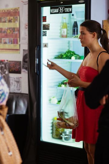Eine Frau in einem roten Kleid steht von einem Kühlschrank mit Glastür und hält eine Plastiktüte mit frischen Lebensmitteln in der Hand.