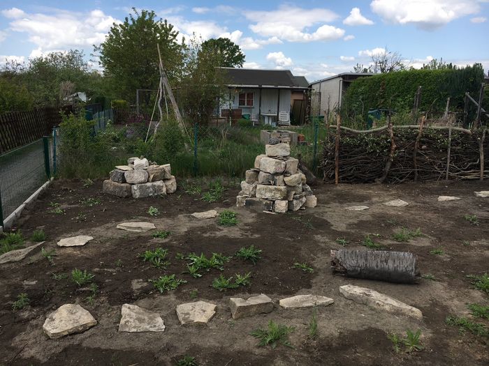 Erste Pflanzen stehen zwischen Wegen aus Trittsteinen in einem Beet. Im Hintergrund sieht man ein Gartenhaus, neben den Beeten steht eine Trockenmauerpyramide.