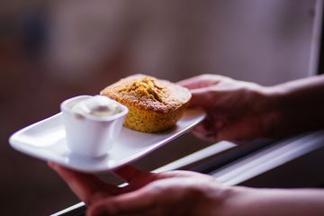 Zwei Hände halten eine kleine Servierplatte, auf der ein heller Muffin und eine weiße Soße angerichtet sind.