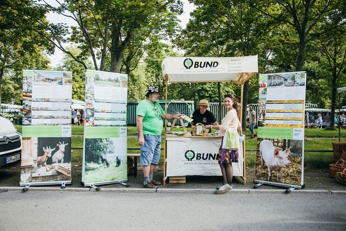 BUND-Stand, neben dem Banner über verschiedene Bauernhöfe rund um Dresden stehen, auf einem Wochenmarkt. Am Stand stehen die Projektreferentin Barbara Braun, das Vorstandsmitglied Gottfried Mann und die Bundesfreiwillige Sabine Förster.  