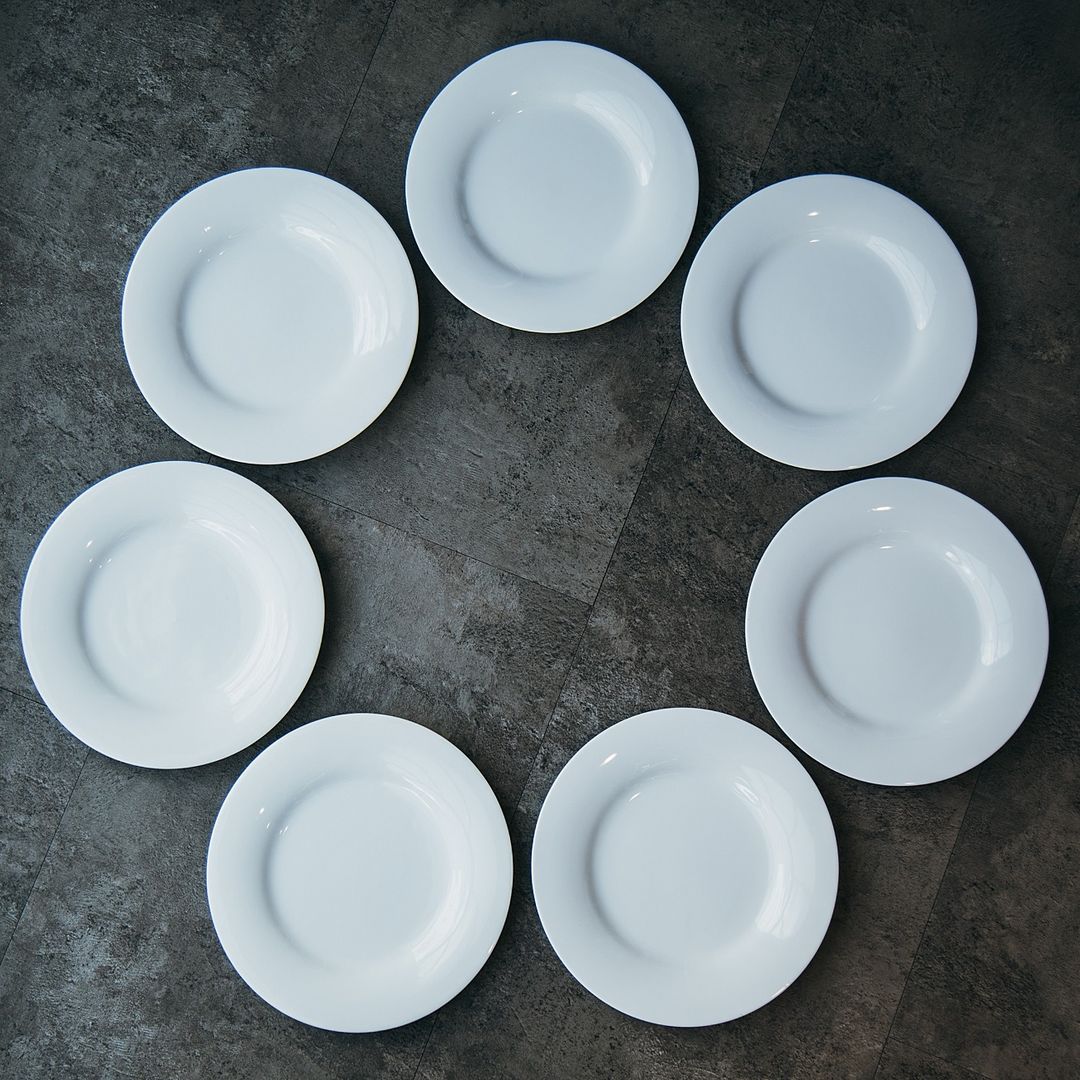 Sieben weiße Teller stehen im Kreis auf dunkelgrauem Untergrund.