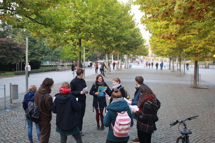 Eine Gruppe von Menschen steht im Kreis auf einem gepflasterten Platz, der von Bäumen umgeben ist.