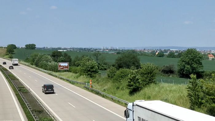 Autos fahren über eine Autobahn, rechts von der Fahrspur steht ein braunes Schild, auf dem Dresden steht und rechts liegt umrandet von etwas Natur die Stadt.