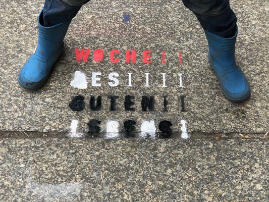 Auf der Straße steht mit ro-schwarz-weißer Sprühkreide geschrieben: Woche des guten Lebens. Darüber steht breitbeinig eine Person mit Gummistiefeln, es sind nur die Beine zu sehen. 