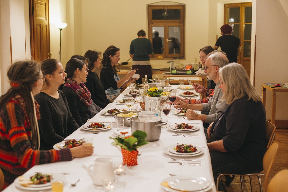 Alle Teilnehmer:innen des Kochkurs sitzen um eine lange, gedeckte Tafel mit weißer Tischdecke und essen ihr selbst zubereitetes Gericht.