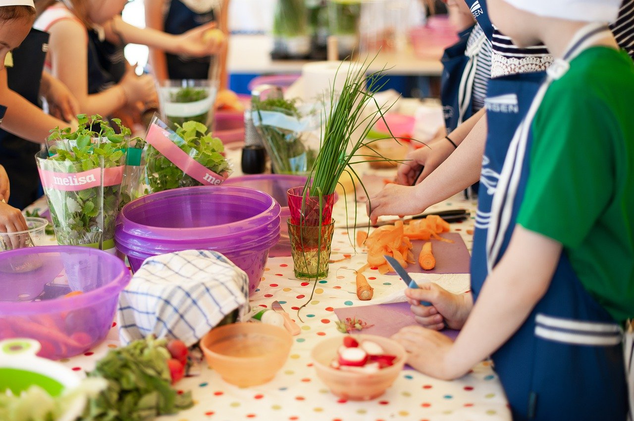 Kinder an einem Tisch, auf dem verschiedene Lebensmittel stehen. Sie schneiden Gemüse.