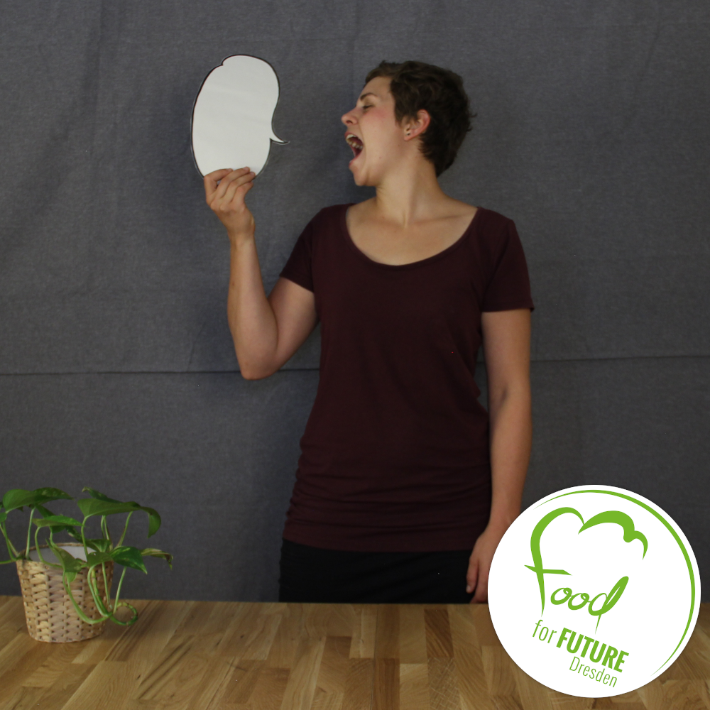 Umweltbildungsreferentin Frederike Becker steht vor dunkelgrauem Hintergrund und hält eine weiße Sprechblase in der Hand, welche sie anschreit. Vor ihr steht ein brauner Tisch, auf dem Kräuter stehen. In der rechten unteren Ecke sieht man das Logo von Food for Future.