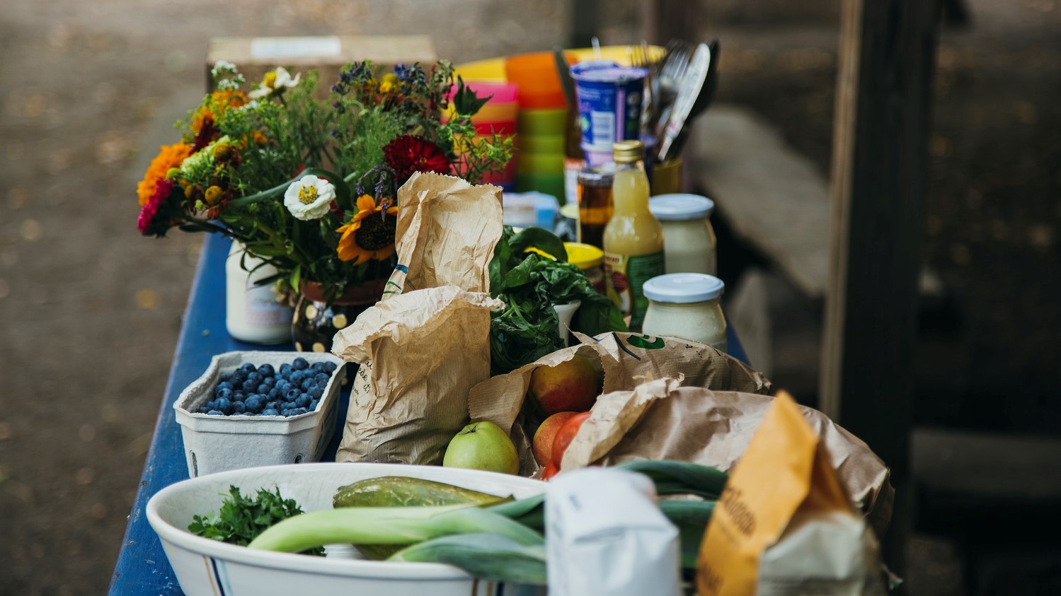 Lebensmittel, die wie frisch vom Einkauf auf einer Küchentheke aufgestellt sind. Darunter ist viel Gemüse und Obst in Pappschalen, sowie ein Blumenstrauß und Milchprodukte in Glasbehältern.