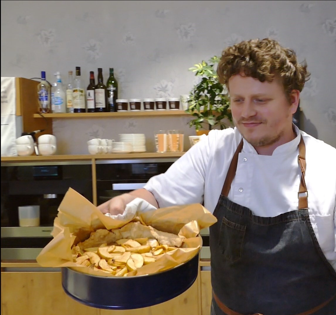 Chef Lutz holds a finished apple pie up to the camera. He is wearing a white T-shirt with a dark blue apron and is standing in a modern kitchen.
