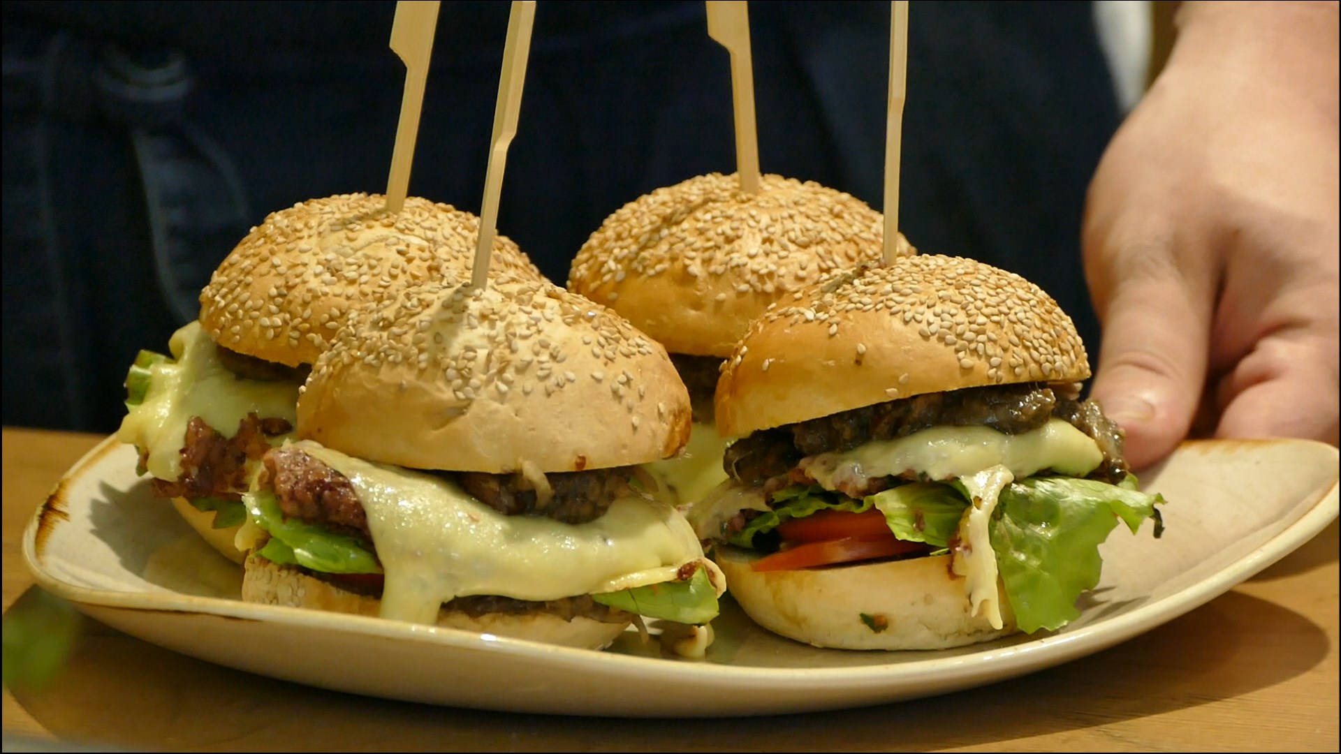 Four small burgers, held together with a wooden stick, are arranged on a plate that is placed on a table by a person.