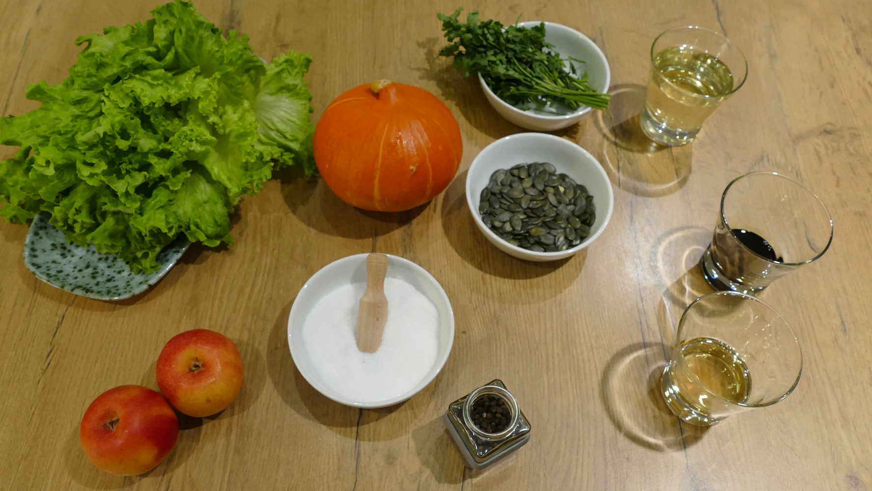 Zutaten wie Salat, Kürbis, Äpfel, Petersilie und Gewürze liegen auf einem hellen Holztisch.