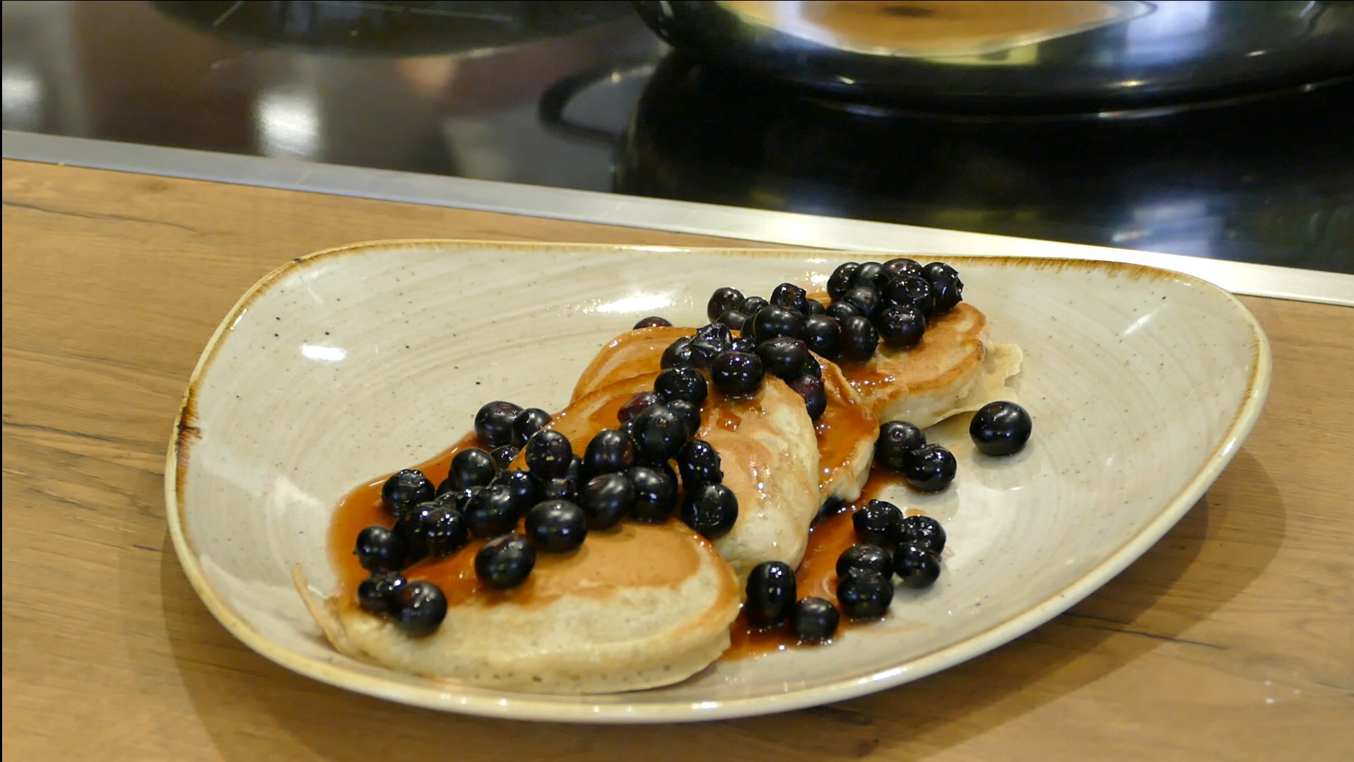 Pancakes are decorated with blueberries on a cream-coloured plate with a golden rim, which is placed on a light wooden table.