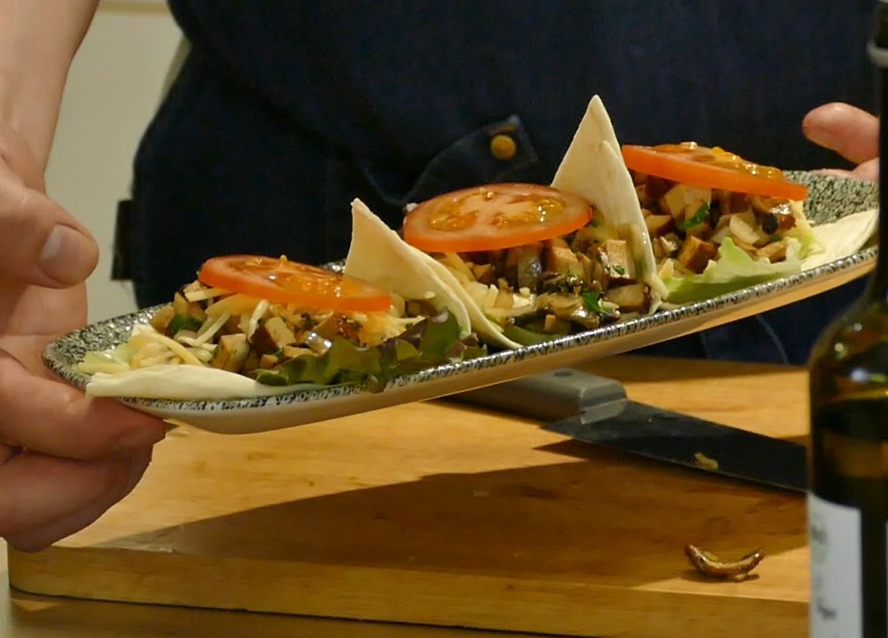 Drei Tacos sind auf einer länglichen Servierplatte angerichtet, die von einer Person in blauem Hemd gehalten wird.