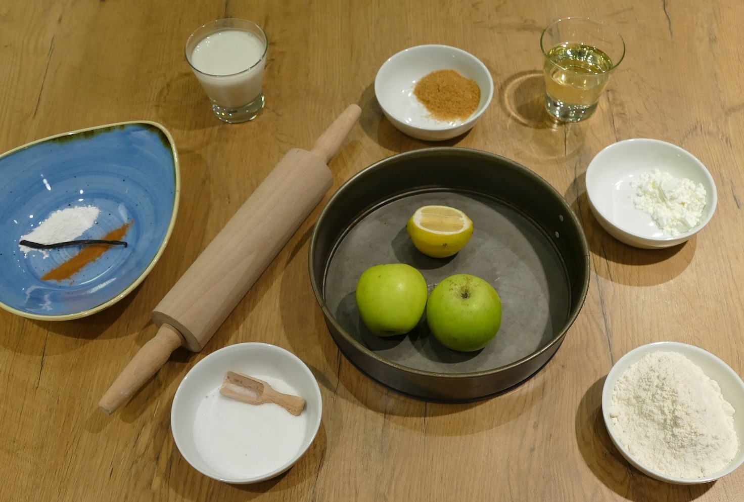 Zutaten wie Äpfel, Vanille, Zucker und Mehl stehen in kleinen Schüsseln angerichtet auf einem Holztisch.