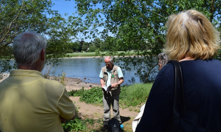 Menschen hören dem Vorstandsmitglied Frank Siegert zu, der vor einem See steht und aus einem Buch liest.