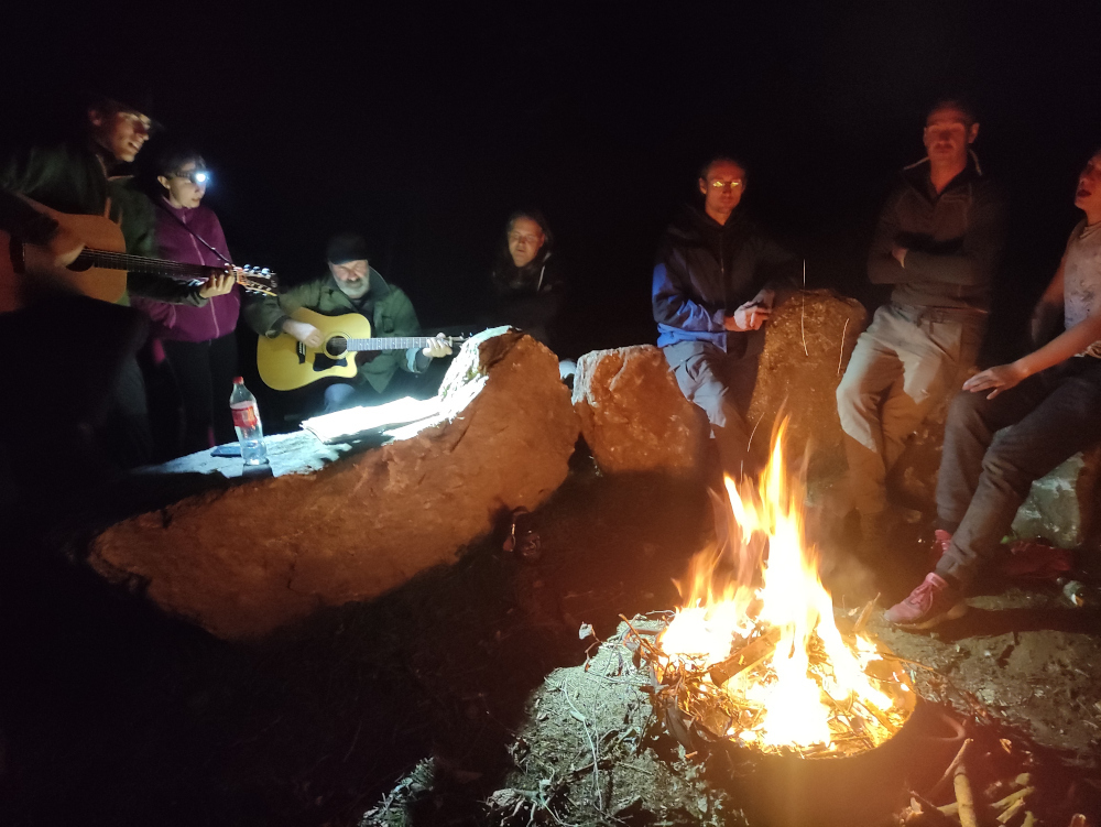 Menschen sitzen auf Steinen um ein Lagerfeuer herum, zwei von ihnen spielen Gitarre.