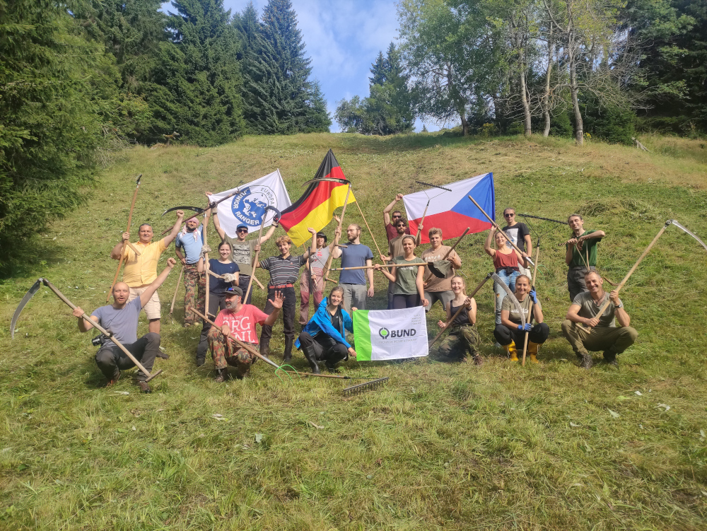 Gruppenbild von Jugendlichen und jungen Erwachsenen, die Arbeitsgeräte wie Sensen oder Rechen in der Hand halten. Hinten halten Menschen die tschechische und die deutsche Fahne nach oben, außerdem werden die Logos der beiden Naturschutzorganisationen in die Kamera gehalten.