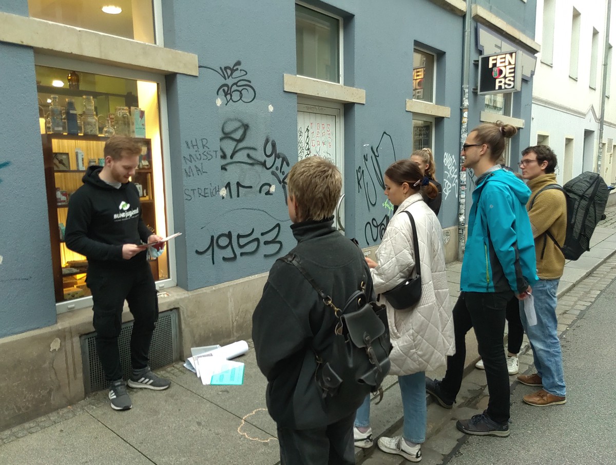 BUNDjugend-Aktiver Marco erzählt ein paar Menschen etwas bei einer Station des konsumbewussten Stadtrundgangs. Sie stehen vor einem blauen Haus.