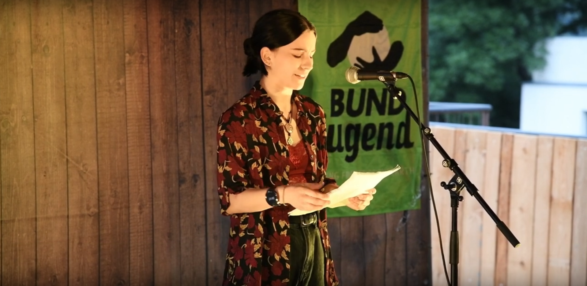 Gewinnerin Sunna Zahn beim Climate-Slam. Sie hält ein Papier in der Hand und spricht ins Mikrofon, hinter ihr hängt eine grüne Fahne der BUNDjugend.