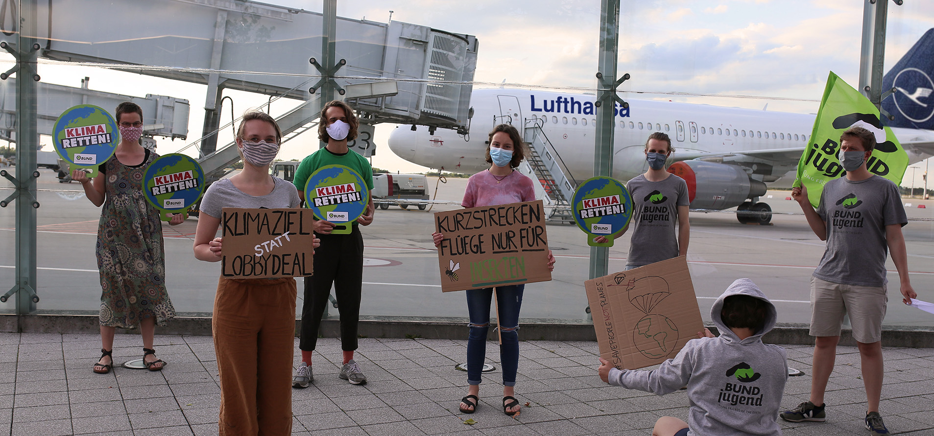 BUNDjugend-Mitglieder am Dresdner Flughafen. Sie tragen Masken und halten Schilder mit den Aufschriften "Klima retten", "Klimaziel statt Lobbydeal" und "Kurzstreckenflüge nur noch für Insekten". 